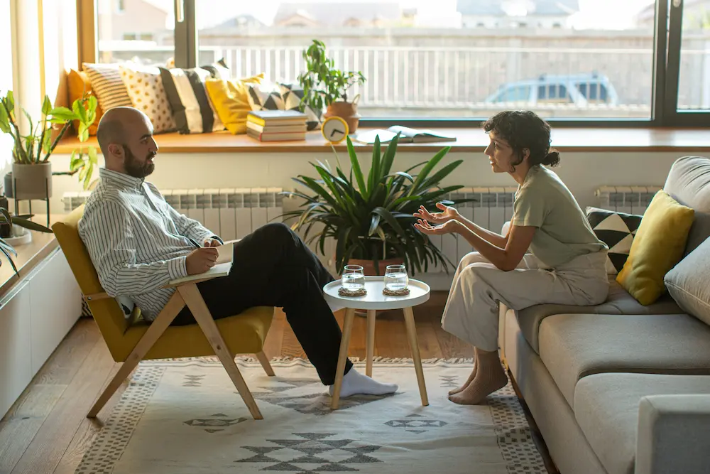 Two people sitting across from each other in a therapy session, talking in a bright room.