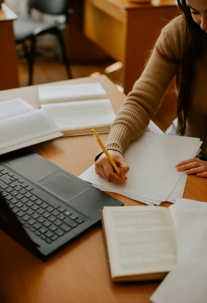 Person writing notes at a desk with a laptop and open books.