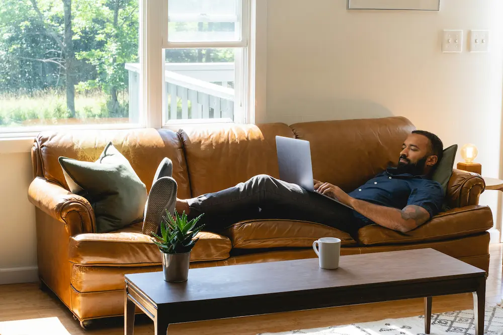 Person working remotely on a laptop while relaxing on a couch at home.